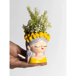 The Citrus Crown Lady Planter is a refreshing decorative piece inspired by sunny Mediterranean aesthetics. Featuring a serene feminine face with closed eyes and soft detailing, this planter is beautifully finished with silver-grey hair and a vivid yellow outfit base. The standout feature is the handcrafted lemon crown circling the rim — detailed with textured yellow lemons and tiny green accents. When filled with greenery or floral arrangements, the plant rises above the crown, creating the illusion of lush natural hair. Citrus Crown Lady Planter